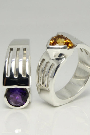 A pair of modern sterling silver bands. One ring features a wide, multi-channel band with a round purple amethyst gemstone, and the other features a tapered band with a triangular amber citrine gemstone.