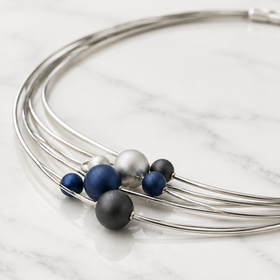 A close-up of a multi-strand silver wire choker necklace with several matte beads in navy blue, charcoal gray, and silver, displayed on a white marble surface 