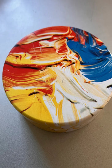 A round decorative box with a colorful abstract paint design in red, orange, yellow, white, and blue.