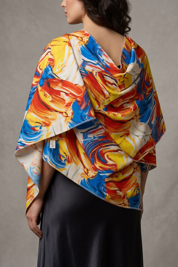A model shown from the back wearing a draped, multi-colored shawl with an abstract paint stroke pattern in blue, yellow, orange, and white over a dark skirt.